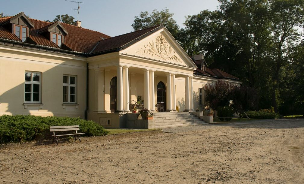 Biały, klasycystyczny dwór z czerwonym dachem i kolumnowym portykiem, otoczony zielenią, przed nim ławka i piaszczysty podjazd