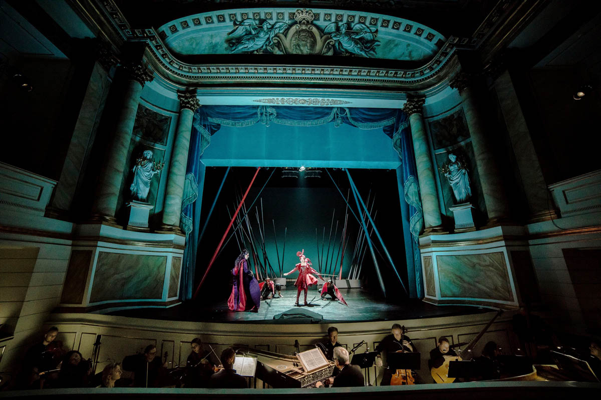 Wnętrze bogato zdobionego, historycznego teatru. Widoczna z dalekiej perspektywy scena ze stojącymi na niej sylwetkami kobiety i mężczyzny.