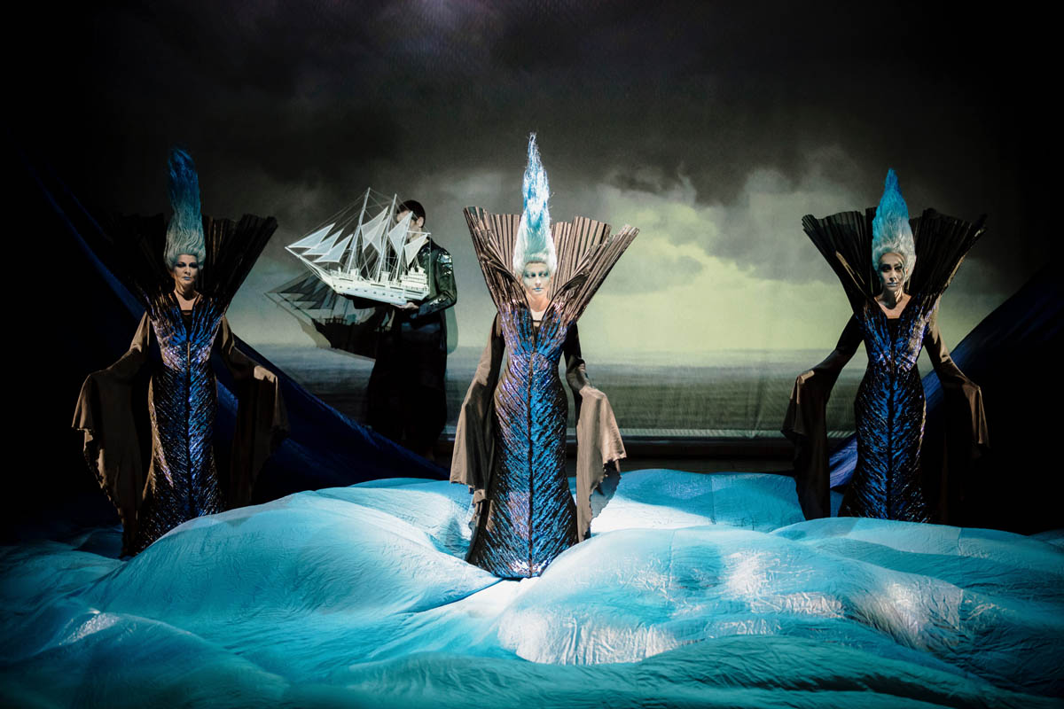 Trzy postacie kobiece w długich sukniach z włosami zaczesanymi wysoko do góry na tle prospektu nieba z chmurami. Stoją na cienkiej, falującej tkaninie, imitującej fale morskie. W tle mężczyzna z małym modelem statku morskiego w dłoniach.