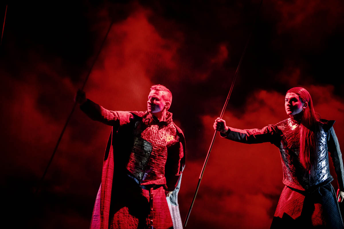 Dwaj mężczyźni stoją obok siebie. Ubrani są w zbroje rycerskie i w peleryny. W dłoniach trzymają cienkie, długie kije.