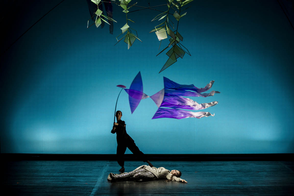 Nad postacią kobiety leżącej na ziemi w długiej sukni stoi postać kobiety w ciemnym obcisłym ubraniu trzymająca w dłoniach długi kij z imitacją lecącego ptaka zrobionego z cienkiej tkaniny. Nad głowami obu postaci zawieszone są formy geometryczne imitujące liście drzewa.