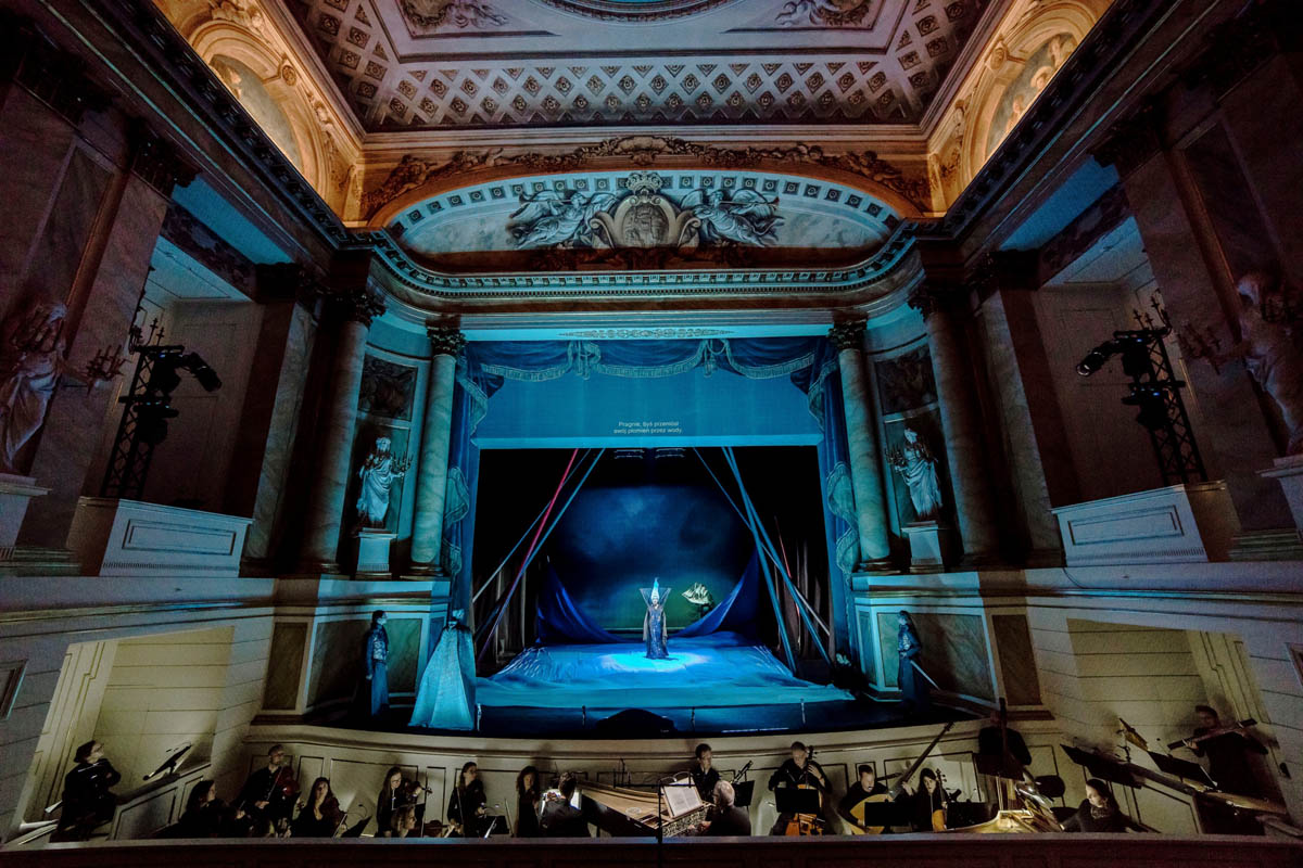 Wnętrze bogato zdobionego, historycznego teatru. Widoczna z dalekiej perspektywy scena ze stojącymi na niej sylwetkami czterech aktorów.