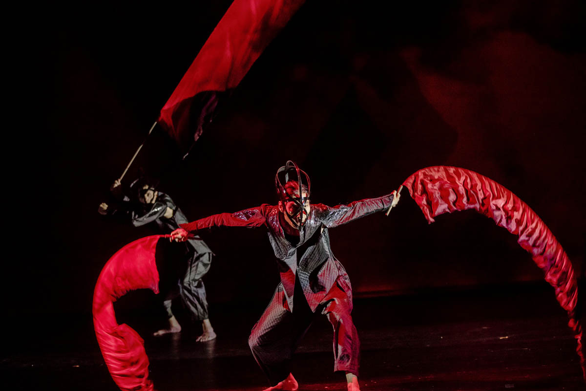 Dwaj mężczyźni w obcisłych strojach poruszają się gwałtownie. W dłoniach trzymają cienkie kije z przytroczonymi do nich pasami cienkiej tkaniny imitującej płomienie ognia.