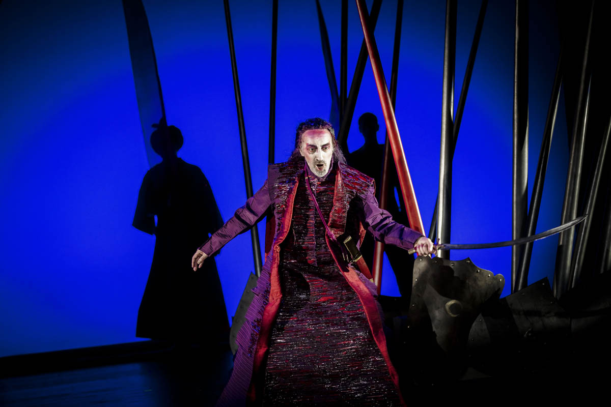 Mężczyzna w długim, bogatym stroju stoi na tle nieregularnie ustawionych wysokich dragów imitujących kopie rycerskie. W tle widoczna leżąca tarcza rycerska i ciemna sylwetka drugiego mężczyzny w dużym nakryciu głowy / turbanie.