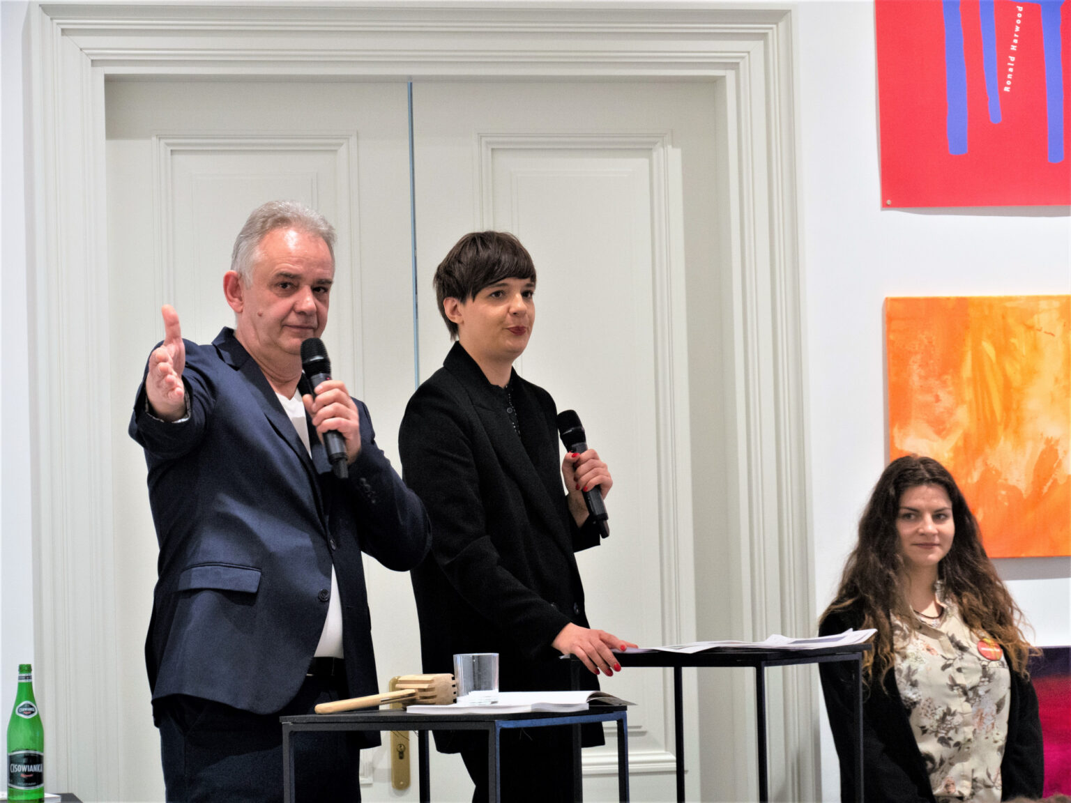 Kobieta i mężczyzna prowadzący aukcję trzymają w rękach mikrofony, mężczyzna wyciągniętą dłonią wskazuje osobę licytującą