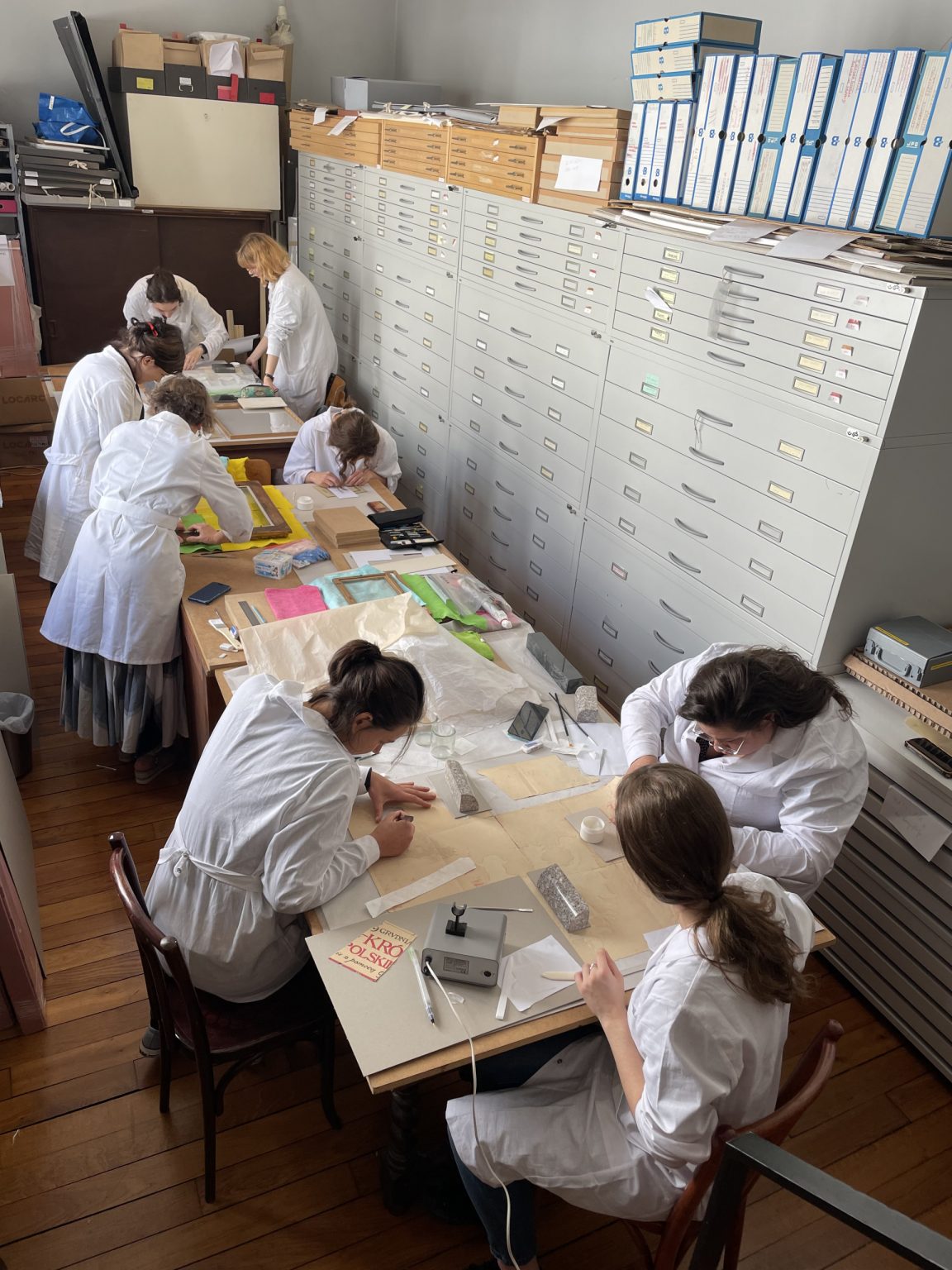 Grupa studentek w białych płaszczach ochronnych wykonuje w pracowni prace konserwacyjne