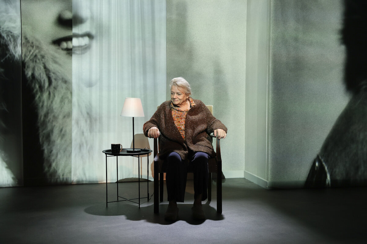 Aktorka siedzi na scenie w fotelu obok niej stolik z zapaloną lampką. w tle wyświetlane z projektora zdjęcie