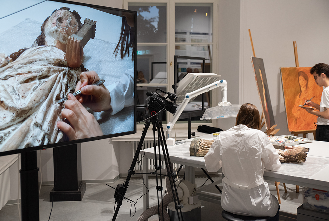 Dwie osoby prowadzące prace konserwatorskie, na ekranie wyświetlane jest zbliżenie wykonywanej pracy