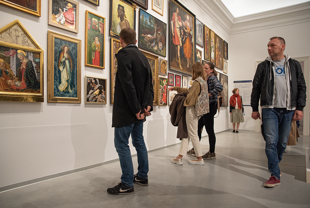 Uczestnicy wernisażu stoją przed eksponowanymi obrazami