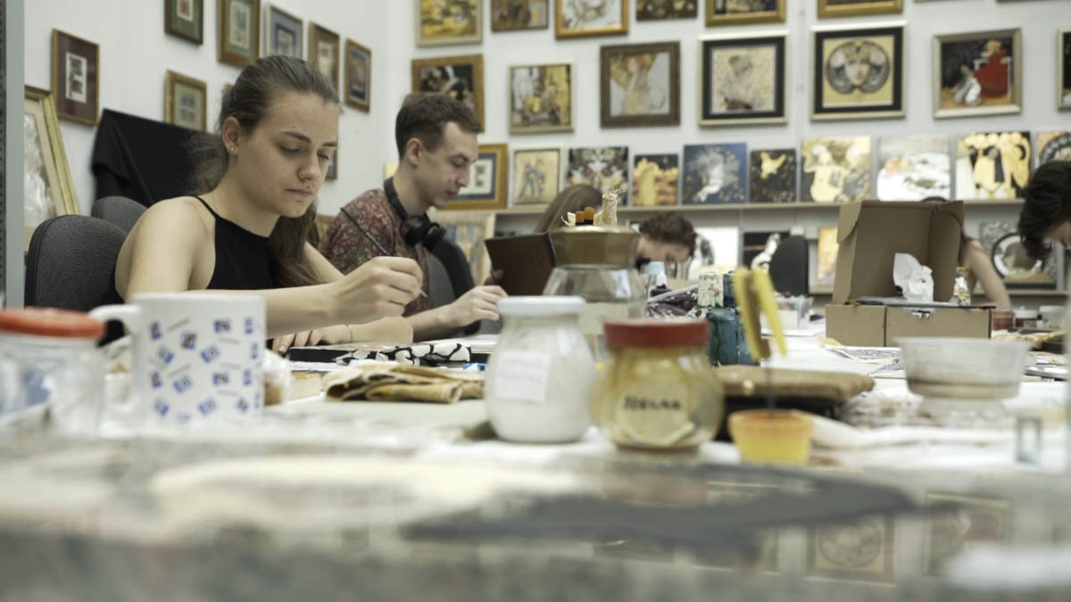 Grupa młodych ludzi siedzi przy stole w pracowni prowadząc drobne prace konserwatorskie