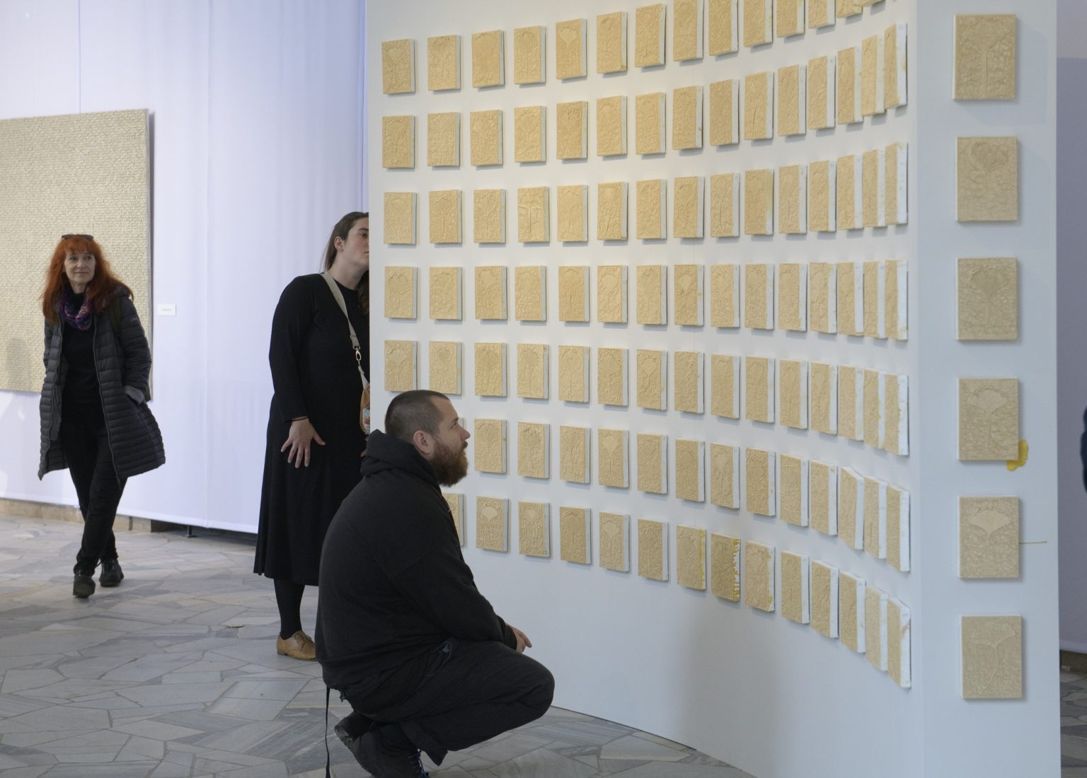 Gość wystawy przed ścianą z zawieszonymi na niej kwadratowymi obrazami o zróżnicowanej fakturze