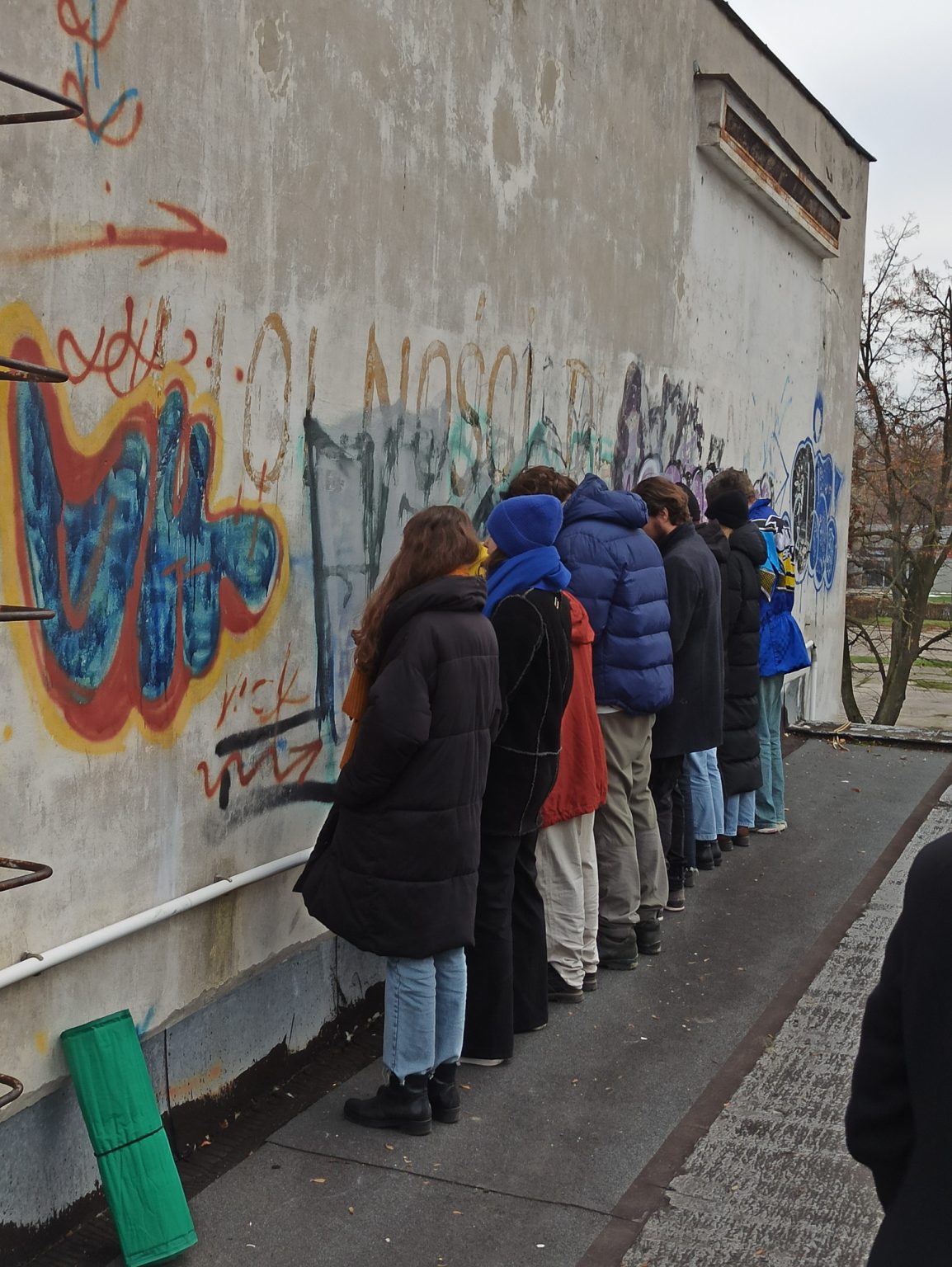 Grupę ludzi stojących w rzędzie twarzami do ściany ozdobionej kolorowym graffiti