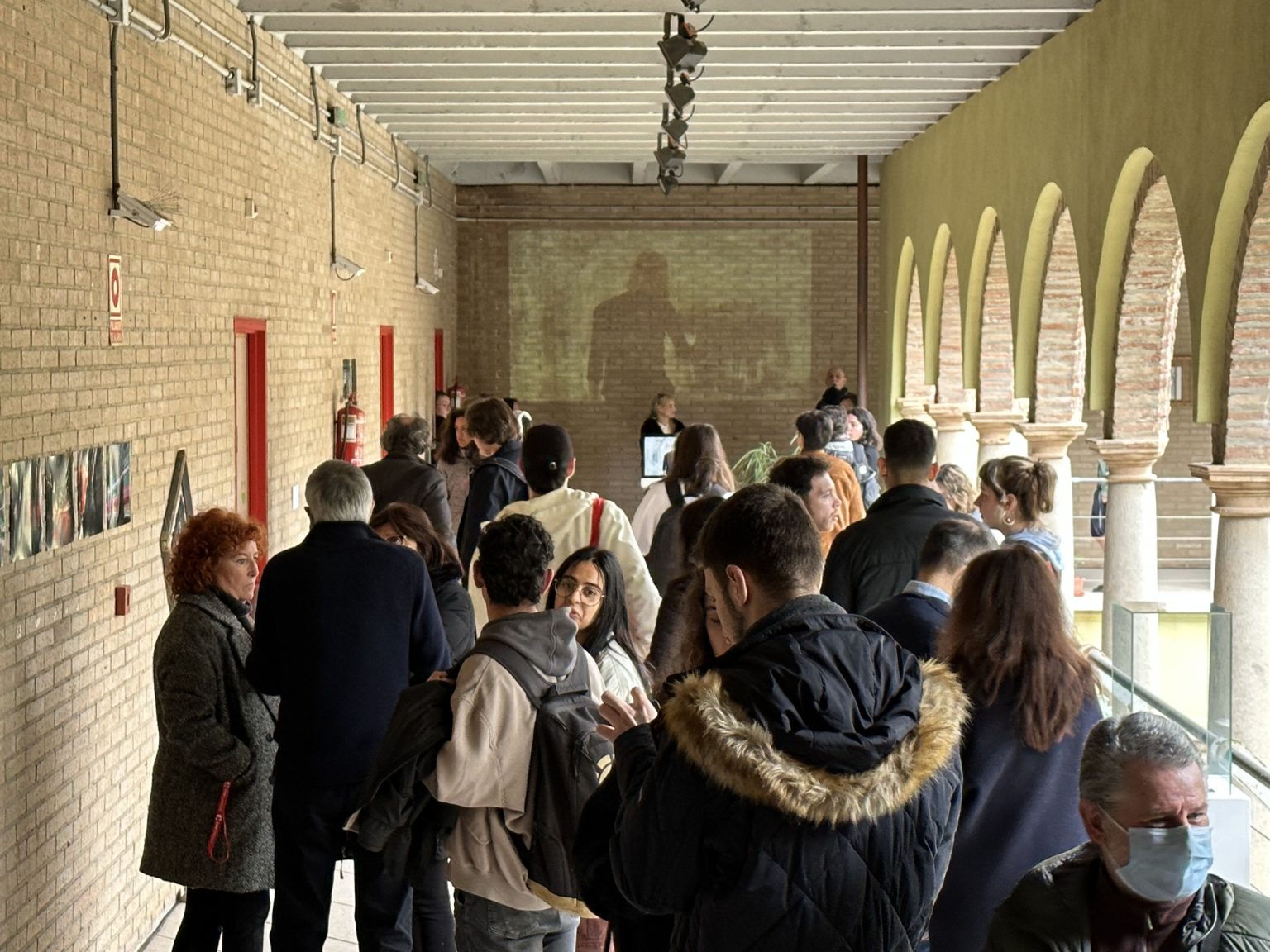 Zwiedzający wystawę na krużganku otaczającym dziedziniec. Na ścianach eksponowane są zdjęcia oraz wyświetlany obraz z projektora