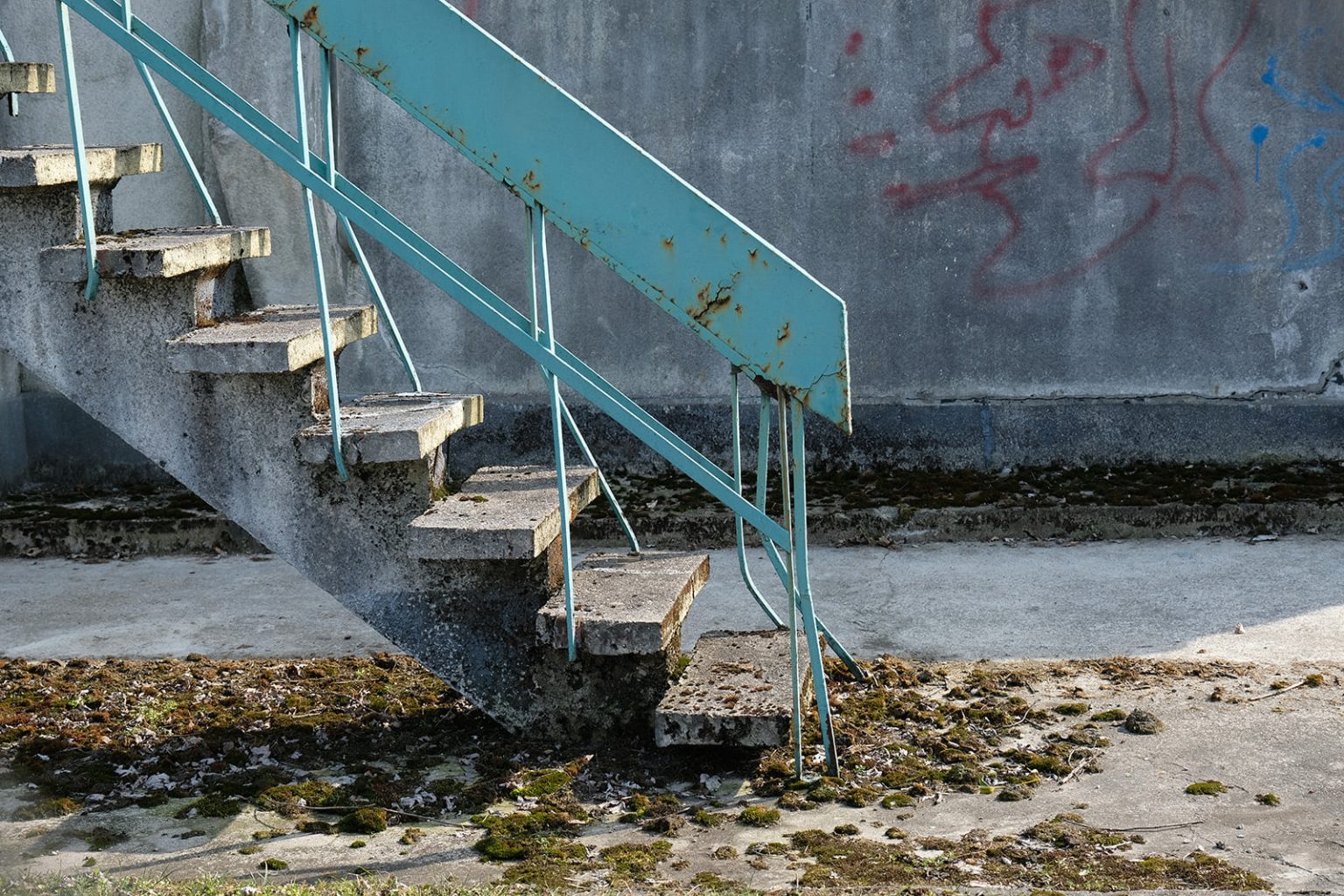 Fragment porośniętych mchem, betonowych schodów. Na pomalowanej na błękitno blaszanej poręczy widać ślady rdzy.