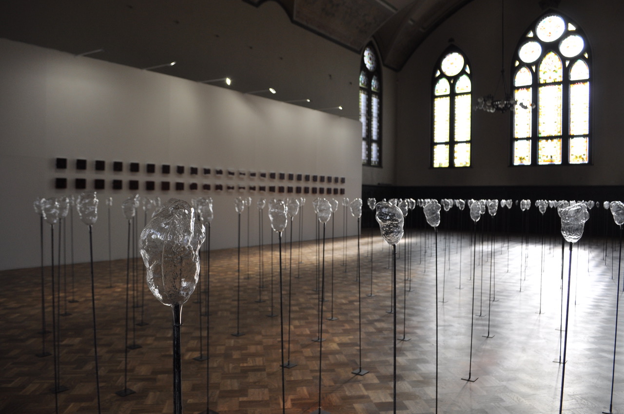 Sala wystawowa wypełniona stojącymi na słupach szklanymi, przezroczystymi rzeźbami, oświetlonymi przez wpadające przez wysokie witrażowe okna światło.
