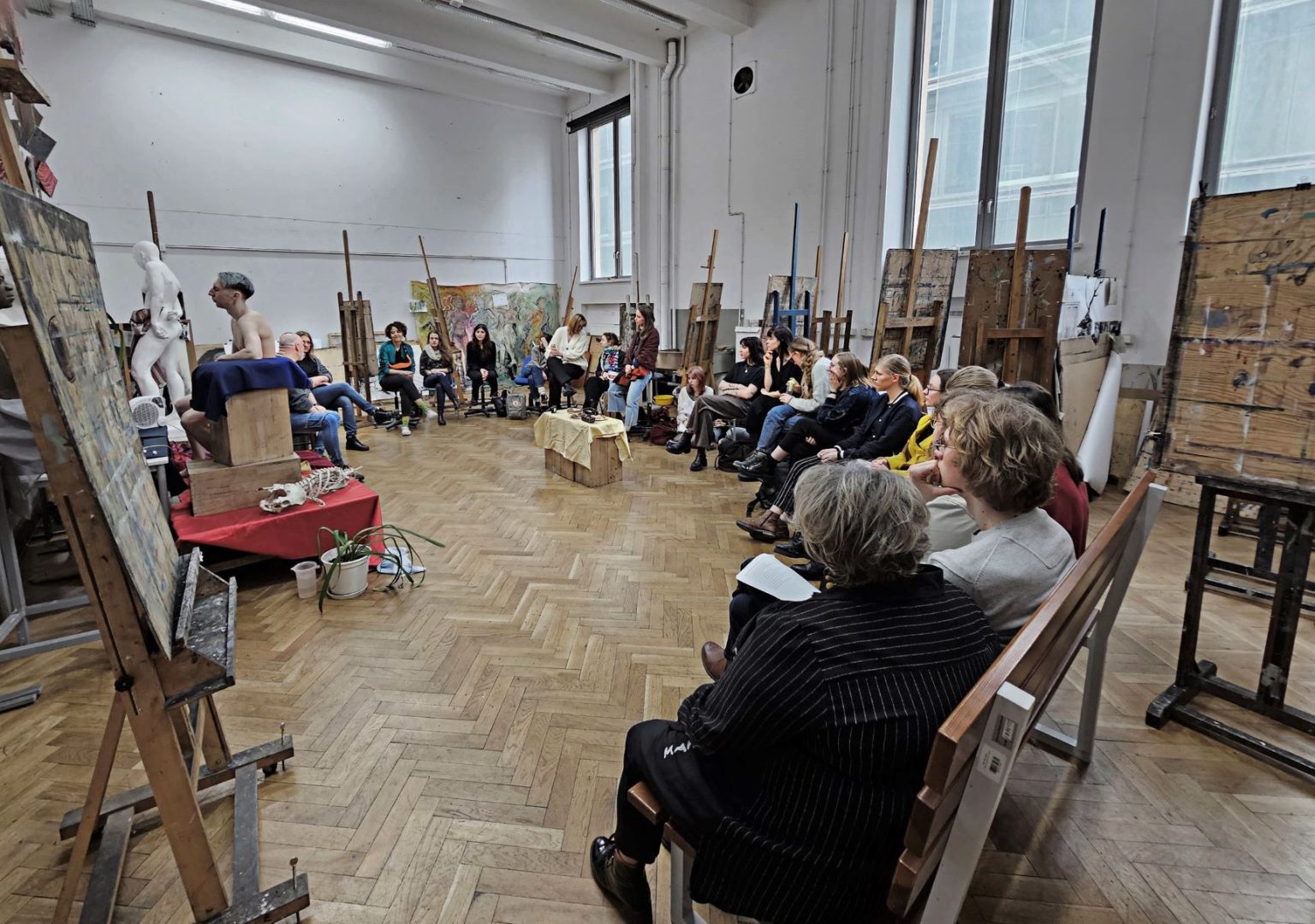 Uczestnicy spotkania siedzą w półokręgu w pracowni. Pod ścianami stoją obrazy na sztalugach
