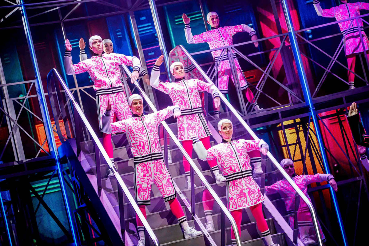 Grupa młodych ludzi wystylizowanych na identyczne plastikowe lalki stoi na tle dekoracji przypominającej wnętrze szkolnej szatni. Ubrani są w jednakowe stroje sportowe i w identycznych nakryciach głowy.