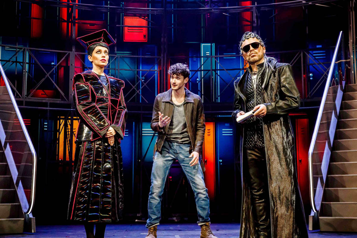 Kobieta i mężczyzna ubrani w błyszczące plastikowe stroje stoją na tle dekoracji teatralnej przedstawiającej szkolne szafki w szatni. Między nimi stoi młody chłopiec ubrany w zwykłe spodnie dżinsowe i kurtkę.