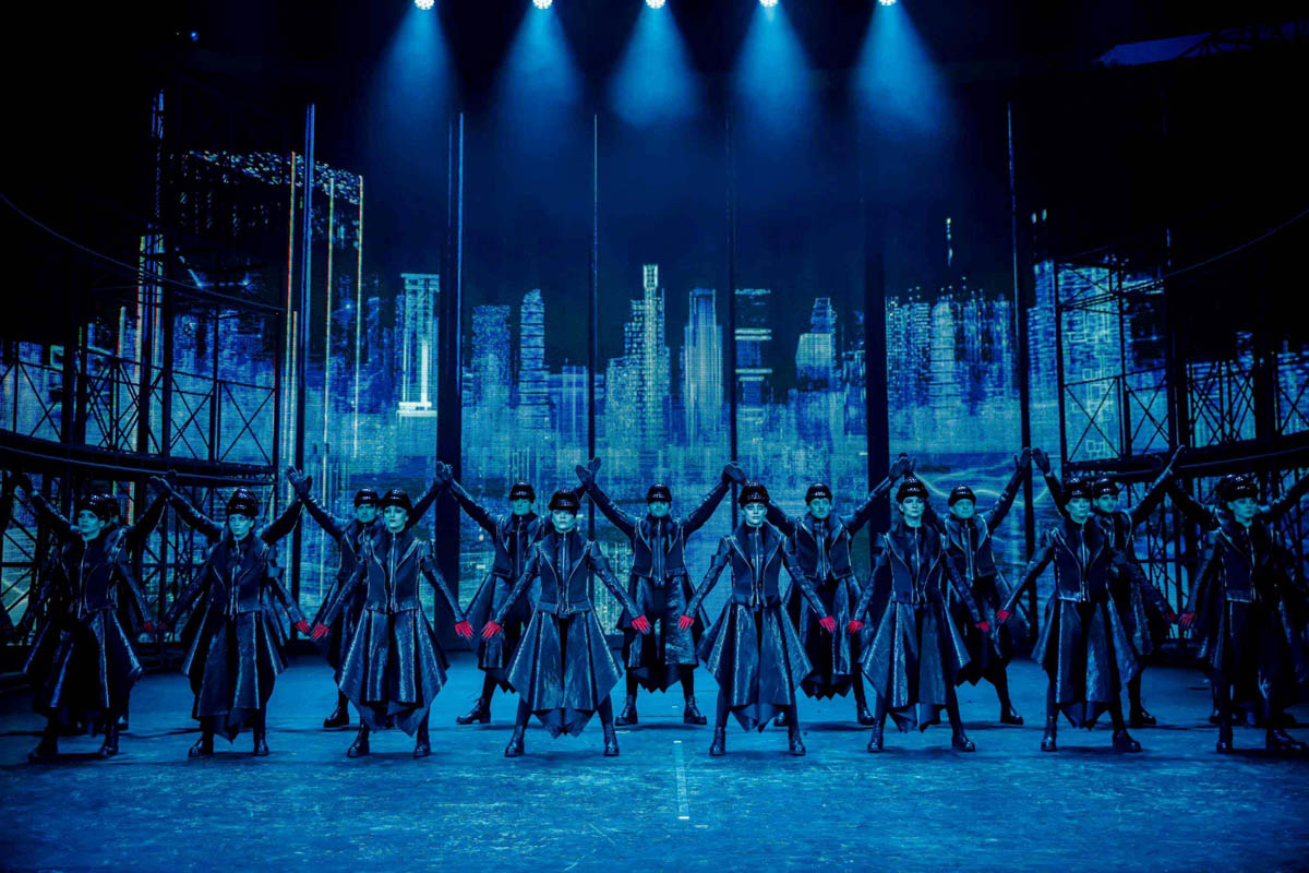 Grupa postaci w błyszczących, ciemnych płaszczach i nakryciach głowy przypominających hełmy stoi w jednakowych pozycjach frontem do widza. Mają naprzemiennie do góry lub do dołu wzniesione lub opuszczone ramiona. W tle dekoracja przedstawiająca wieżowce współczesnego, wielkiego miasta w nocy.