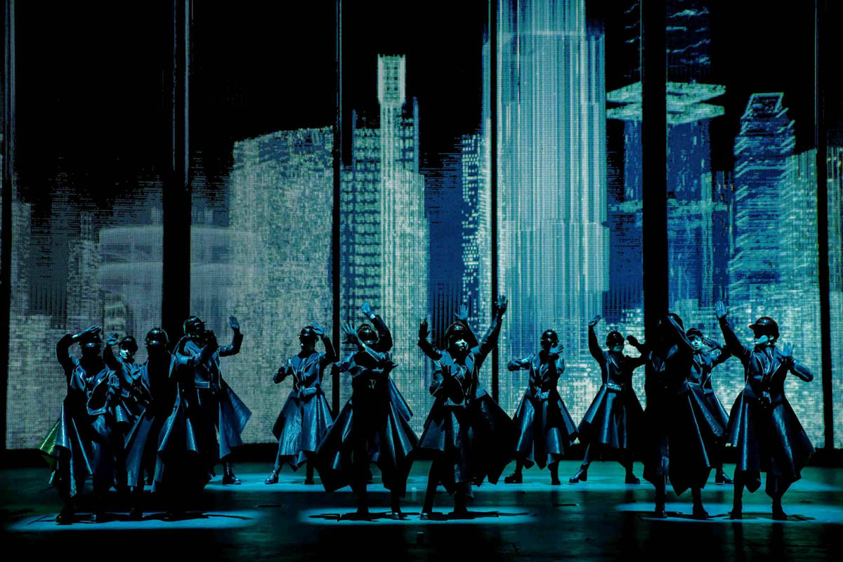 Grupa ciemnych postaci w szerokich płaszczach i w nakryciach głowy przypominających hełmy. Stoją w rożnych dynamicznych pozach na tle dekoracji przedstawiającej współczesne wieżowce wielkiego miasta w nocy.