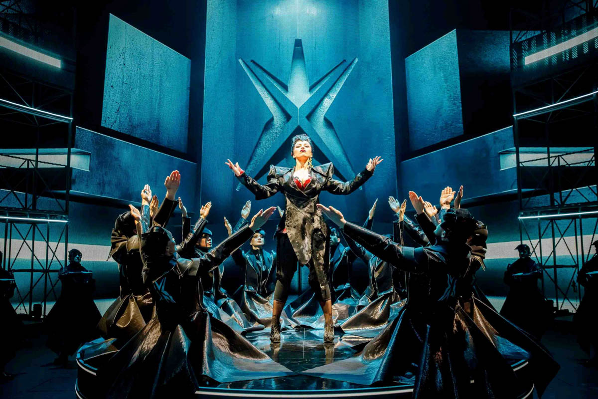 Grupa postaci ubranych w błyszczące, ciemne płaszcze klęczy z poniesionymi ramionami wokół stojącej w centrum kobiety ubranej w strój kojarzący się z dyrektorką współczesnej organizacji korporacyjnej. Stoją na tle dekoracji przedstawiającej betonowe wnętrze nowoczesnego biurowca.