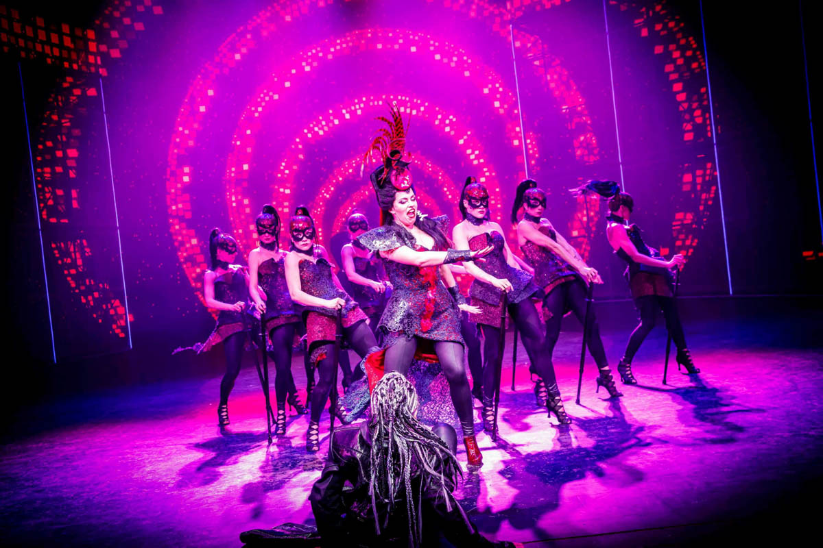 Tańcząca grupa dziewcząt ubranych w efektowne błyszczące, krótkie sukienki i fantazyjne nakrycia głowy.