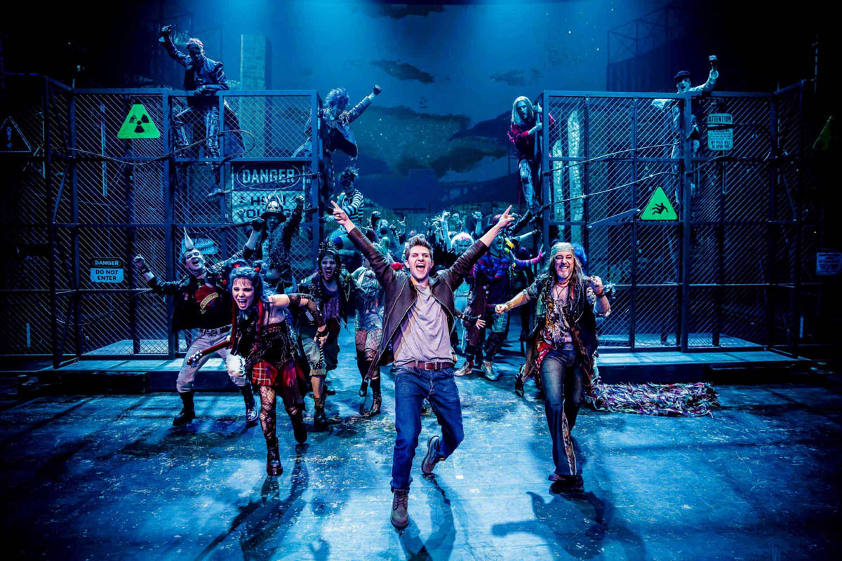 Duża grupa biegnących do przodu uśmiechniętych młodych ludzi ubranych w stroje nawiązujące do sposobu ubierania się gwiazd muzyki rockowej lat 70-tych i 80-tych. W tle dekoracja teatralna przedstawiająca ogrodzenie stadionu sportowego z metalowej siatki po której się wspina kilka postaci.