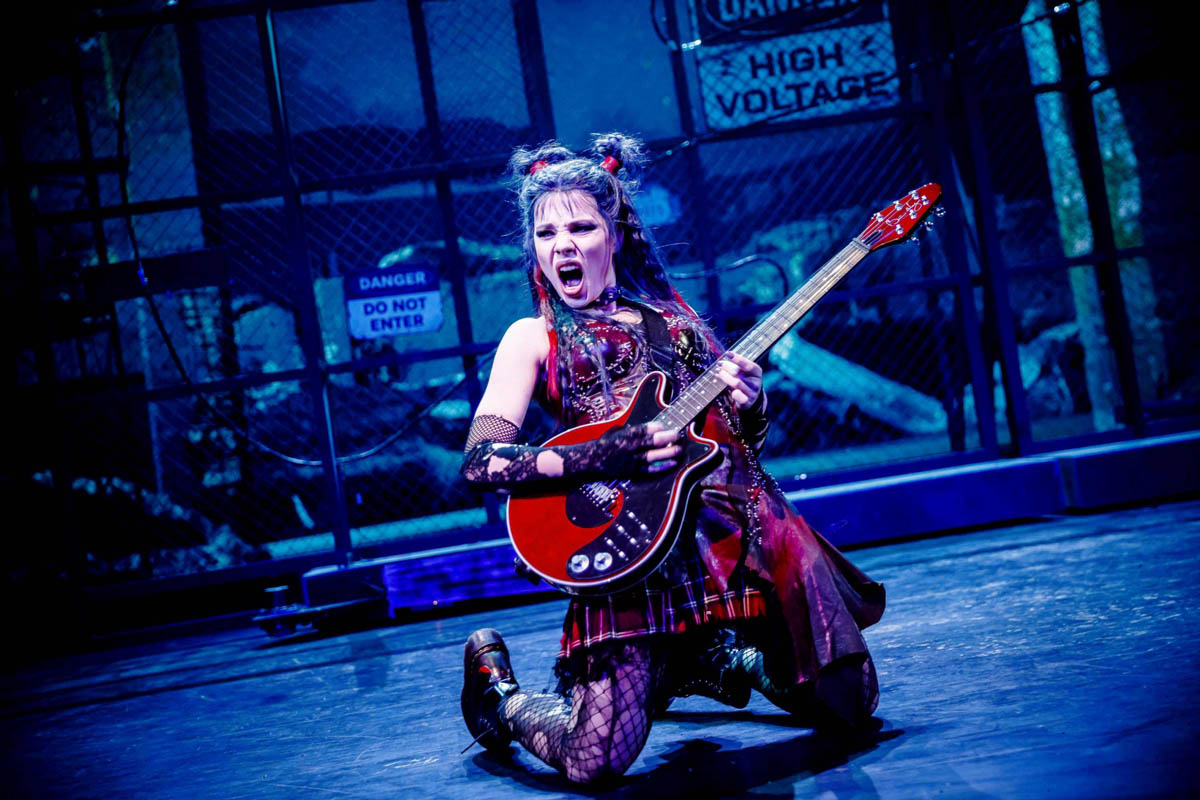 Młoda kobieta ubrana w strój nawiązujący do stylu subkultur młodzieżowych - skórzany gorset, krótką spódniczkę i ciężkie sznurowane buty - gra na gitarze i śpiewa w pozycji klęczącej. W tle dekoracja przedstawiająca ogrodzenie stadionu sportowego z metalowej siatki.