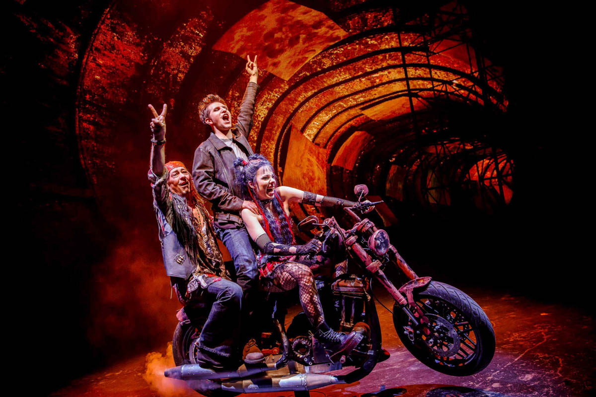 Dziewczyna i dwóch mężczyzn z uniesionymi do górę ramionami i z palcami dłoni ułożonymi w symbol zwycięstwa jadą na dużym motorze. Dziewczyna prowadzi motor, który sprawia wrażenie rozpędzonego w biegu. W tle dekoracja przedstawiająca opuszczony tunel metra.