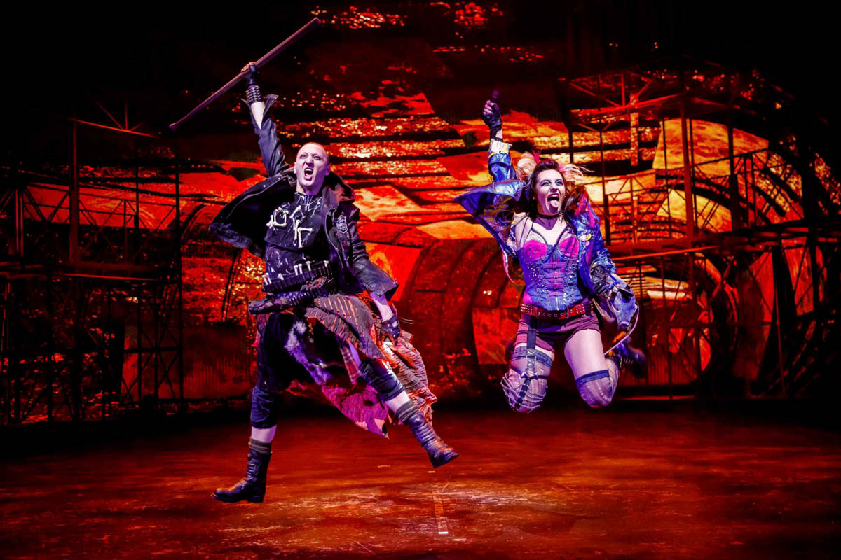 Dziewczyna i chłopak skaczą do góry z uniesionymi ramionami. Ubrani są w pełne fantazji stroje nawiązujące do sposobu ubierania się gwiazd estrady związanych z muzyką rockową. W tle dekoracja przedstawiająca wnętrze opuszczonej stacji metra.