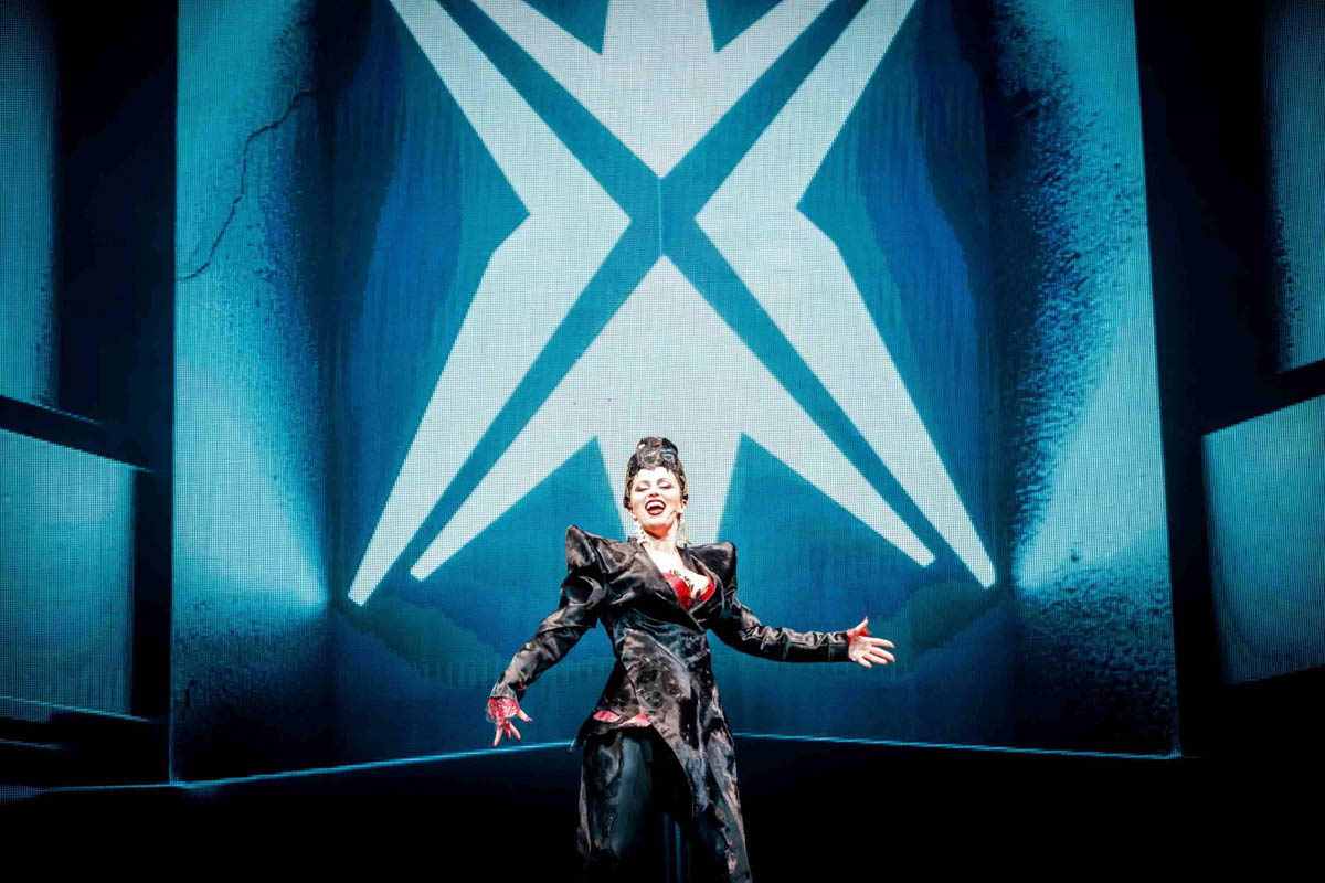 Uśmiechnięta kobieta stoi na tle ogromnego znaku graficznego symbolizującego współczesną wielką organizację korporacyjną. Ubrana jest w ciemny strój z bardzo szerokimi ramionami nawiązujący do stylu ubierania się współczesnych dyrektorek wielkich biur.