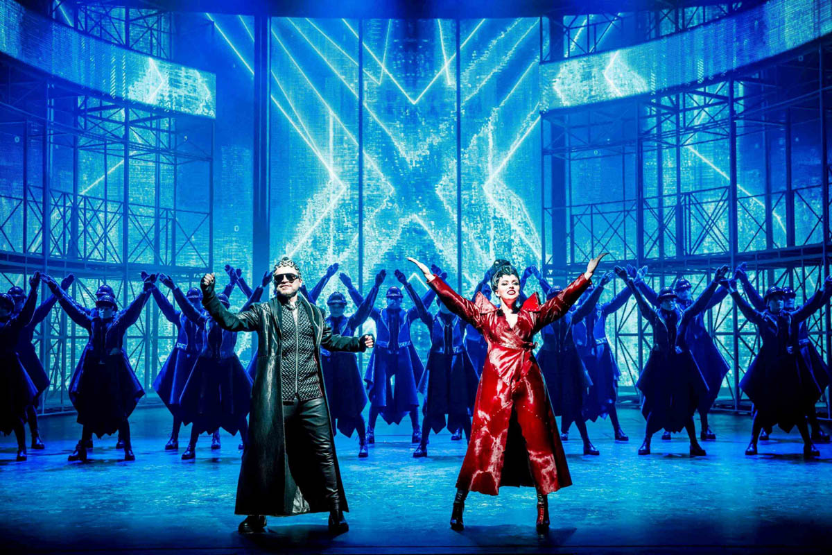 Kobieta i mężczyzna w dekoracyjnych, błyszczących długich płaszczach stoją z uniesionymi do góry rękoma. Za nimi widać sylwetki postaci w ciemnych szerokich płaszczach stojące na tle metalowych rusztowań.