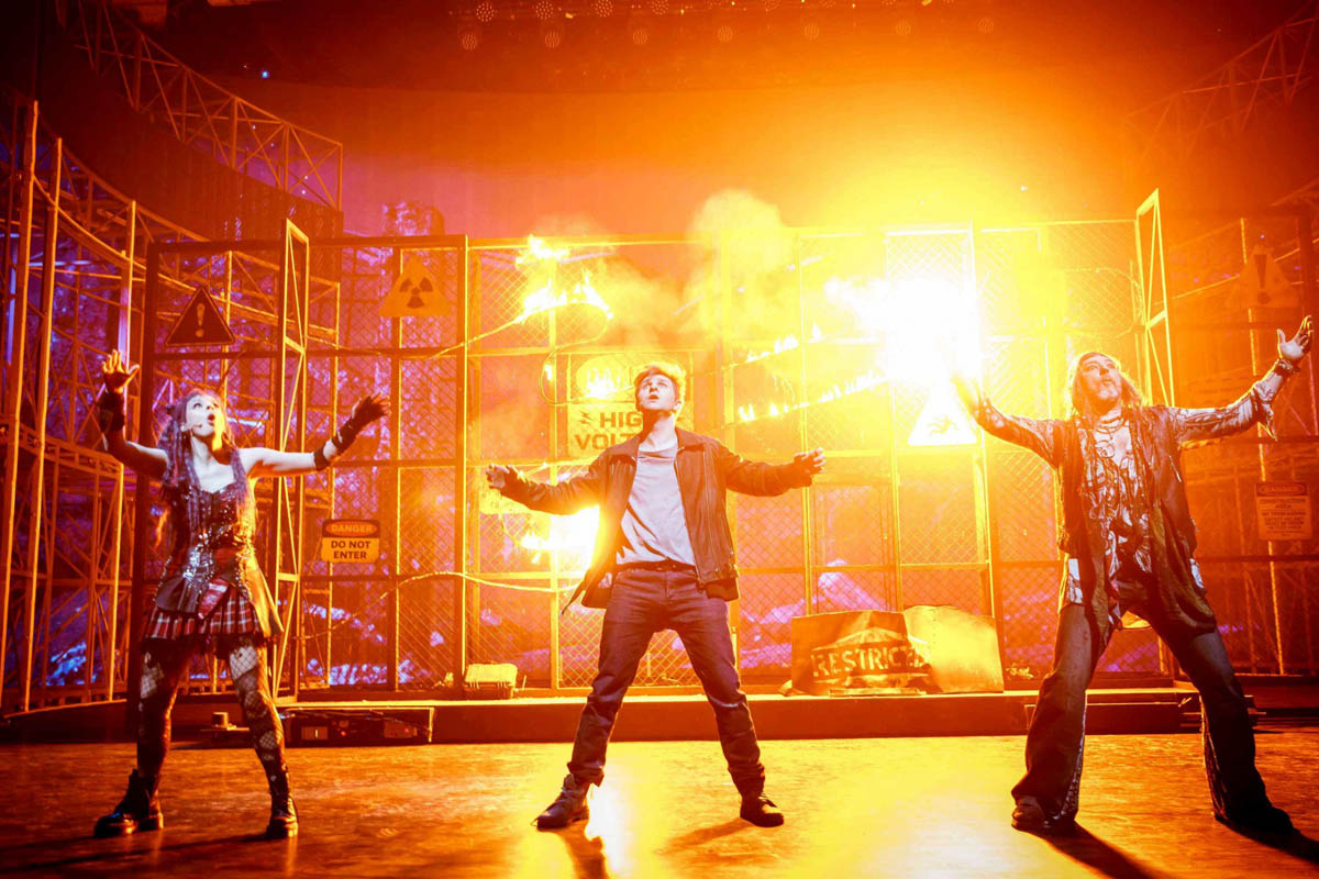 Kobieta i dwóch mężczyzn ubranych we współczesne, młodzieżowe stroje stoją na tle dekoracji przedstawiającej ogrodzenie stadionu sportowego z metalowej siatki. Robią wrażenie przestraszonych. Za siatką widoczne wybuchy z ognia.