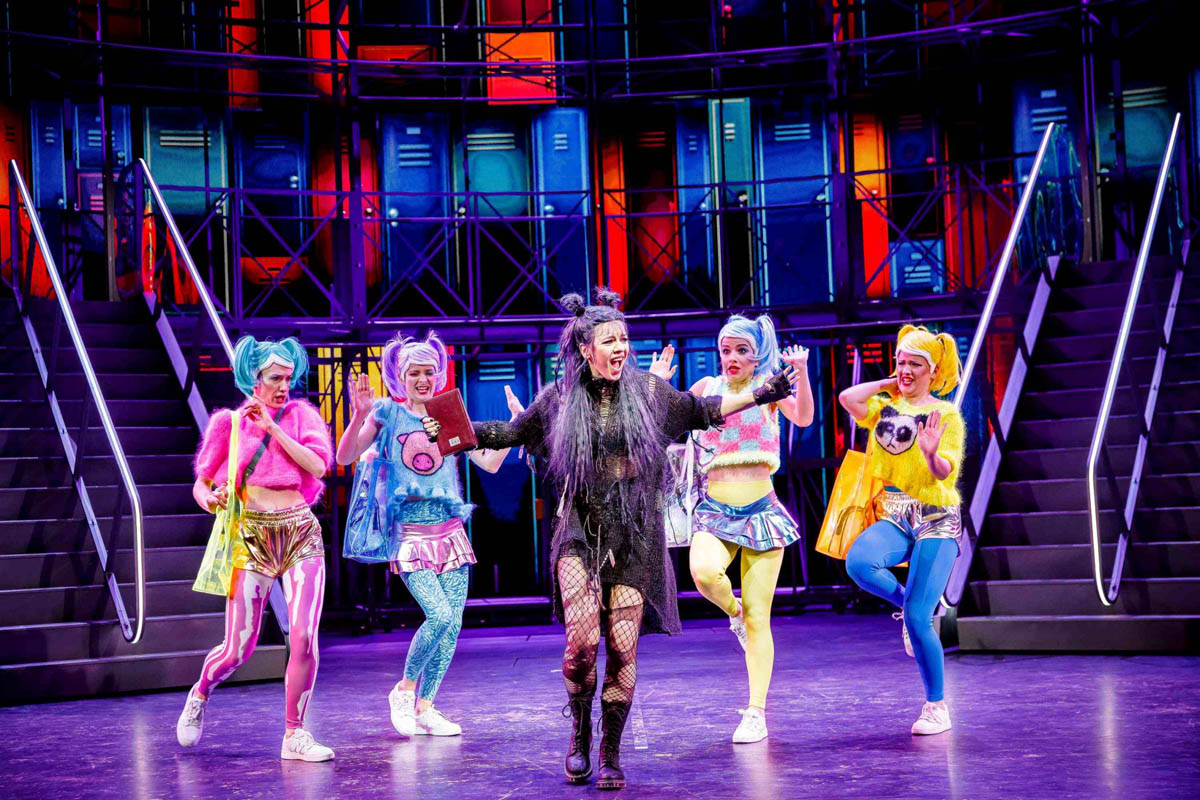 Pięć młodych kobiet w dynamicznych pozach ciała stoi na tle dekoracji przedstawiającej wnętrze szkoły. Ubrane są w zabawne stroje nawiązujące do stylu ubierania się współczesnej młodzieży.