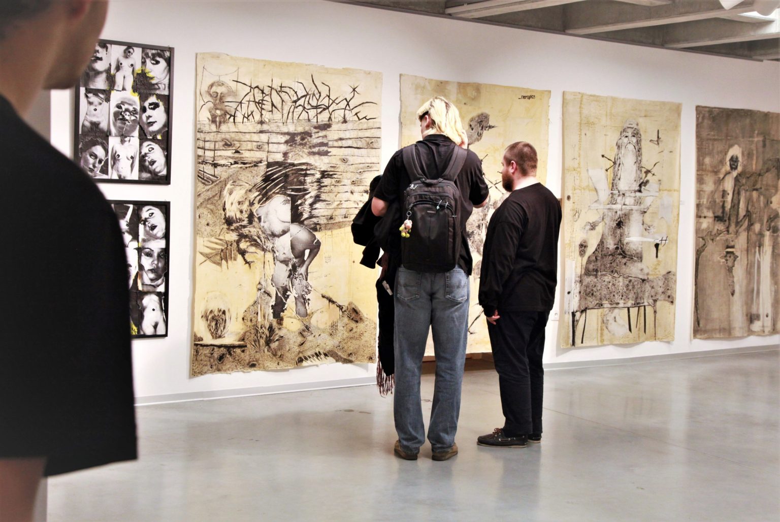 Dwóch mężczyzn w przestrzeni wystawowej. Na ścianie eksponowany jest kolaż, utworzonych z czarno-białych zdjęć