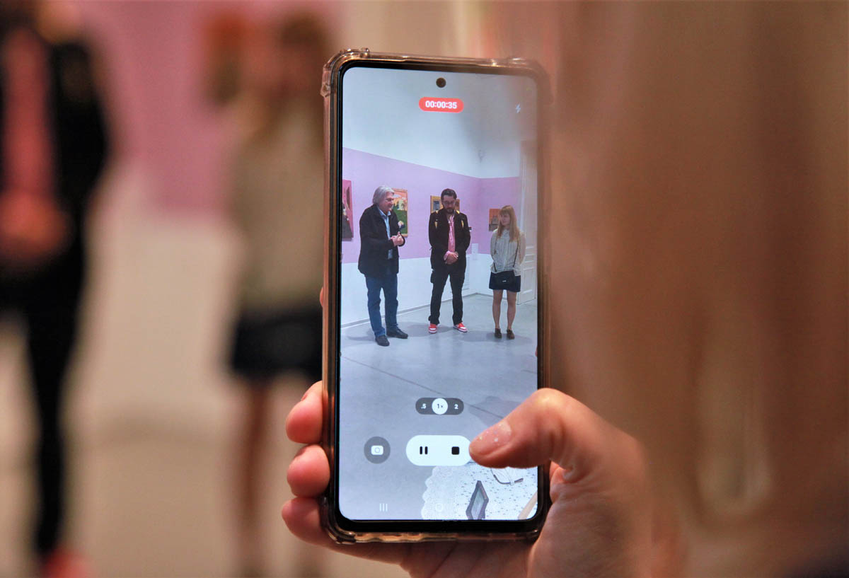 Zbliżenie na dłoń trzymającą działający w trybie nagrywania telefon, na którego ekranie widać grupę 3 osób przed białą ścianą z zawieszonymi na niej obrazami.