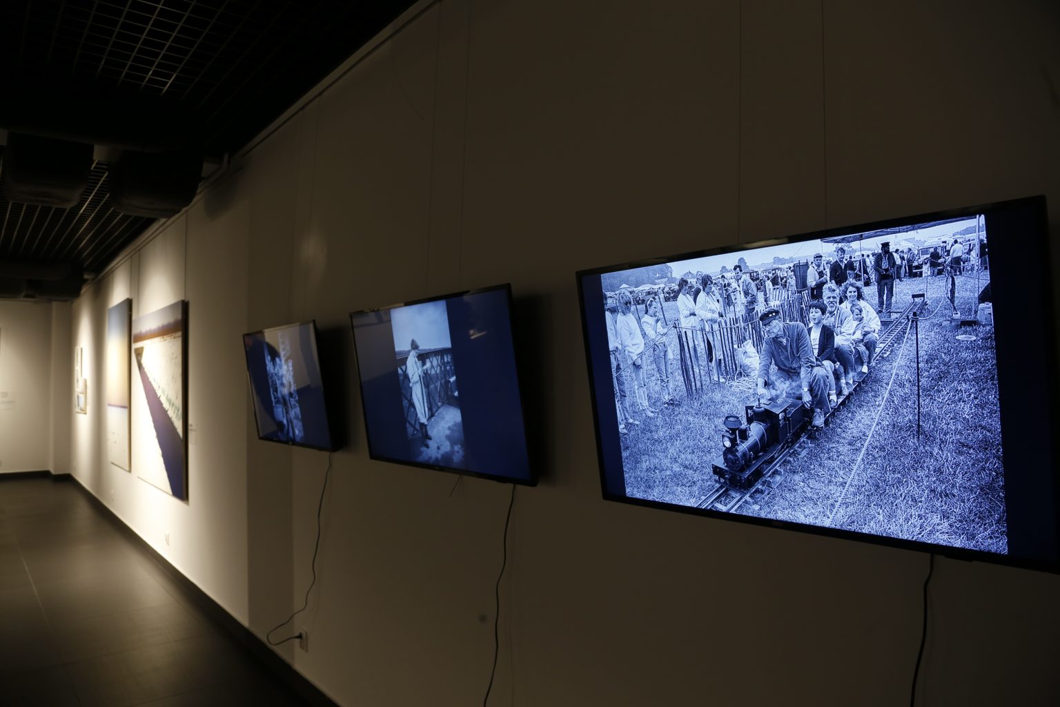 Przestrzeń wystawoa. Na zawieszonych na ścianie monitorach wyświetlają się czarno-białe fotografie.