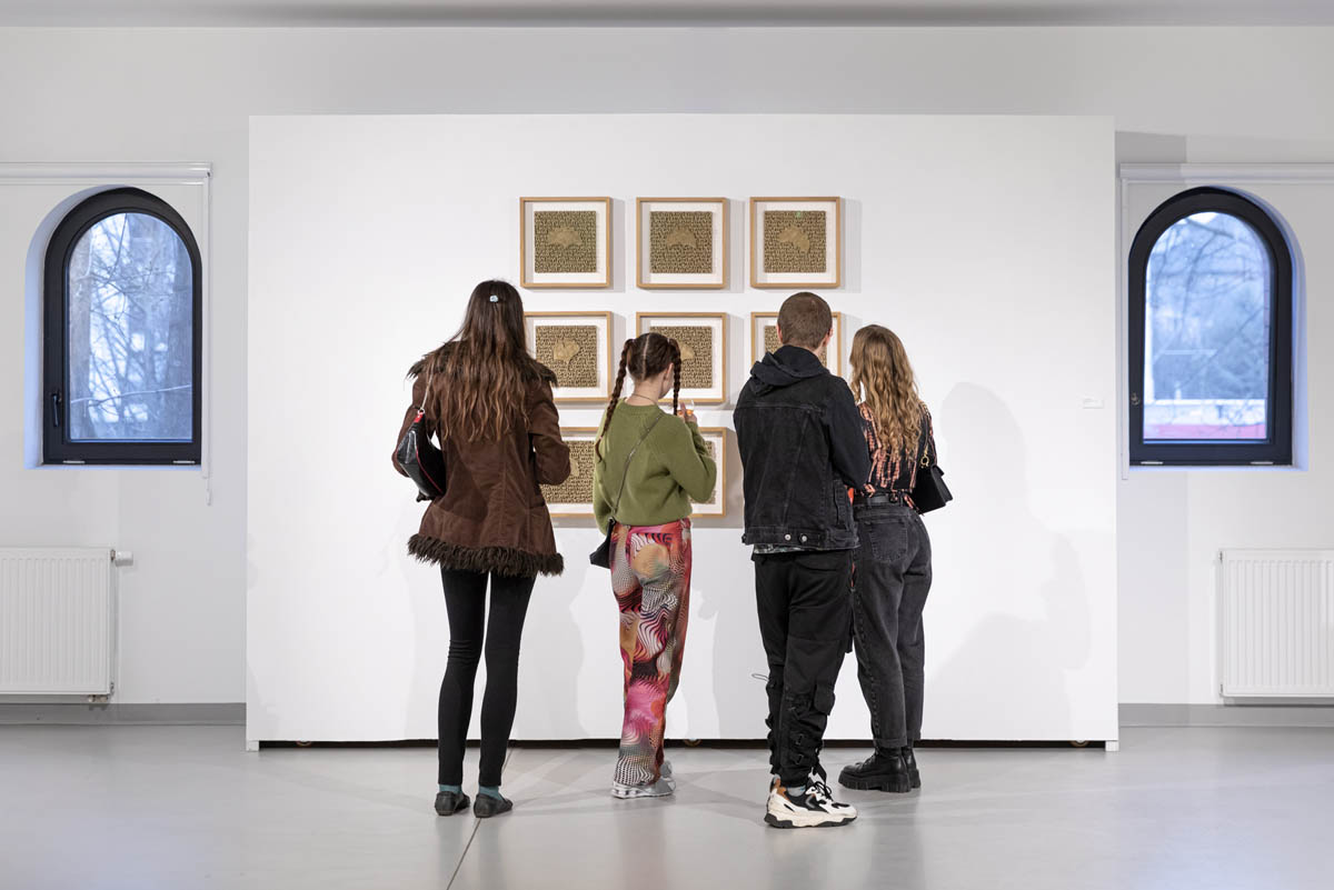 Grupa osób stoi przed eksponowanymi na ścianie pracami.