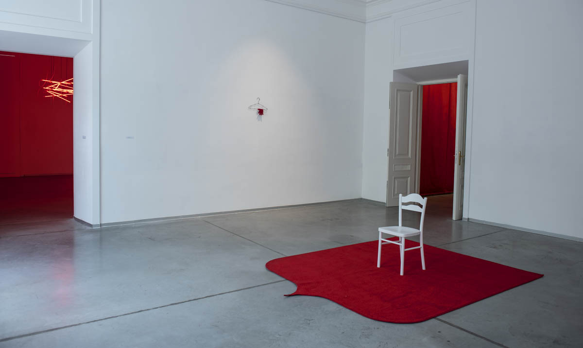 Białe krzesło postawione na jaskrawo czerwonym dywanie. Dywan dodaje koloru wnętrzu, kontrastując z dominującą szarością przedniej i tylnej części planu.