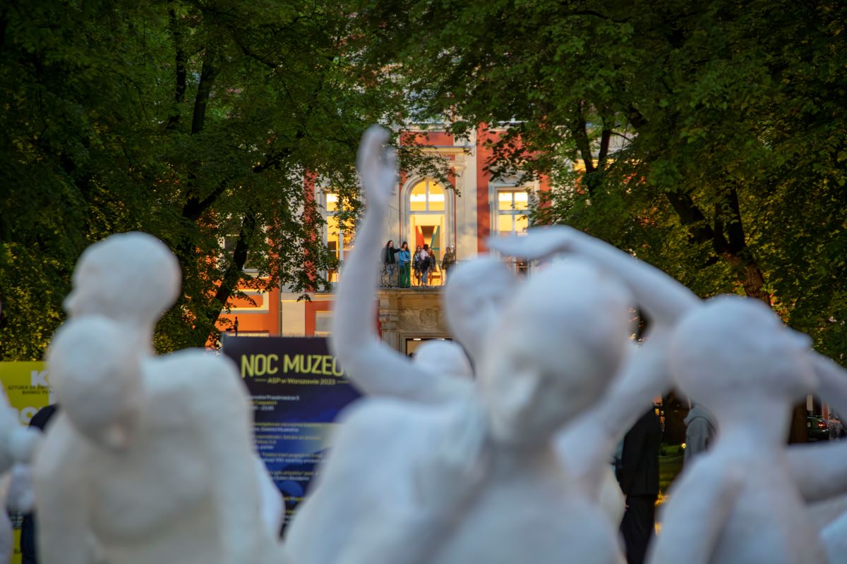 Oświetlona fasada Pałacu Czapskich widoczna spomiędzy rzeźb postaci ludzkich ustawionych na dziedzińcu