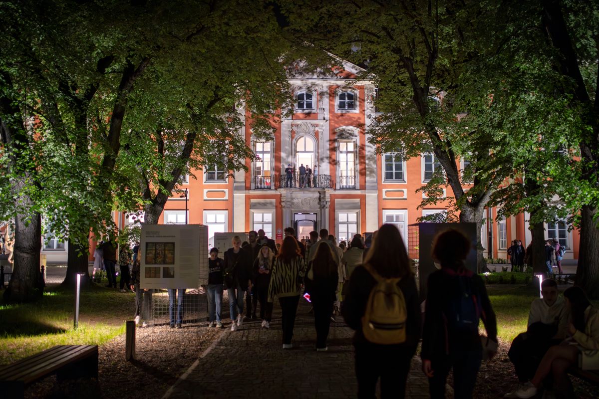 Zdjęcie nocne grupy ludzi na dziedzińcu przed podświetlonym budynkiem Pałacu Czapskich