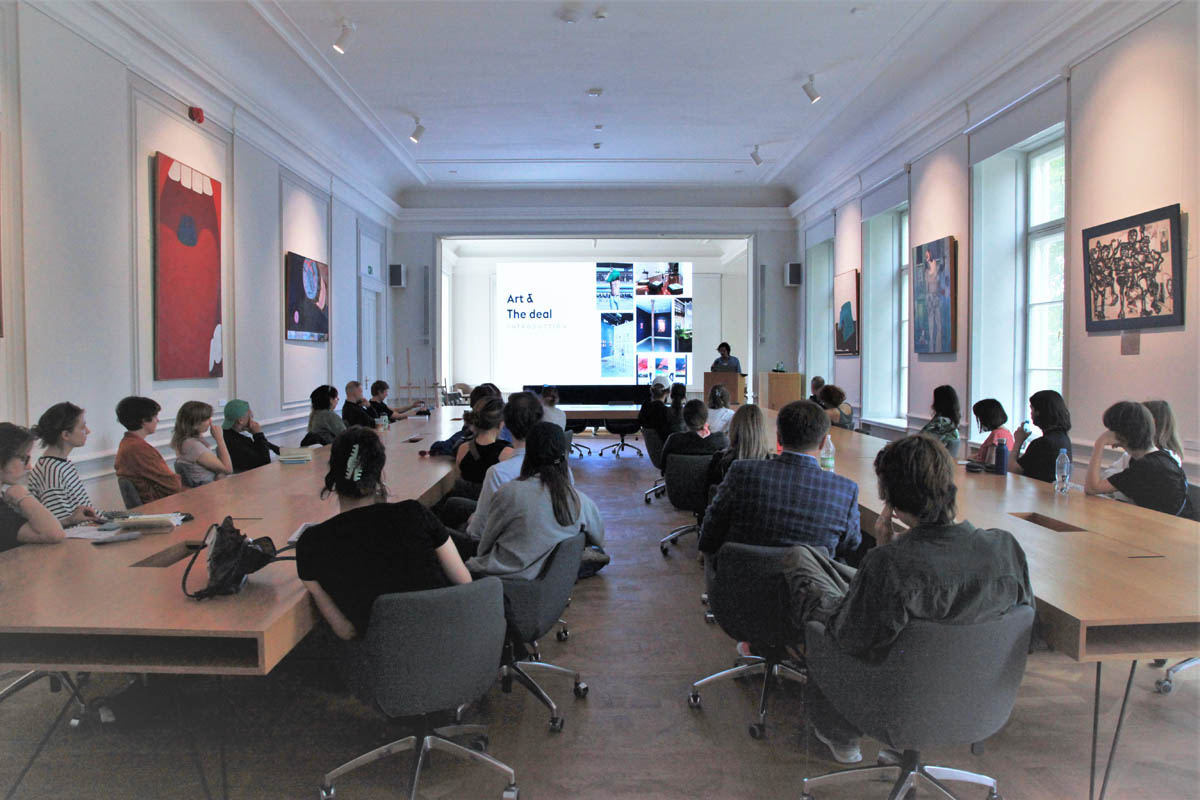 Publiczność podczas wykładu. W tle za mownicą prelegent, za nim slajd z kolażem zdjęć i hasłem "Art & The deal"