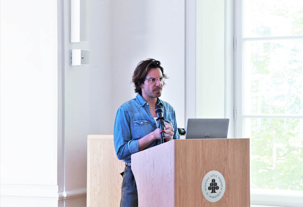 Alex Mor przemawia w Sali Senatu ASP. Stoi za mównicą z naniesionym logo ASP.