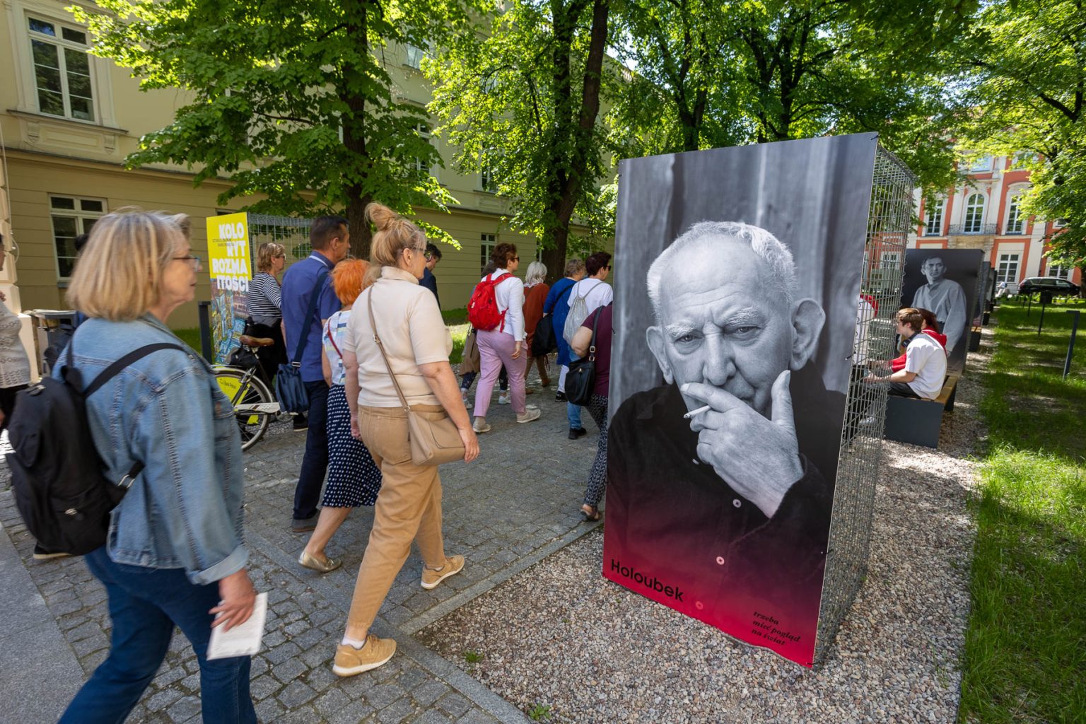 Spacerujący ludzie wchodzą na dziedziniec Pałacu Czapskich. Po obu stronach chodnika stoją duże reprodukcje zdjęć Gustawa Holoubka