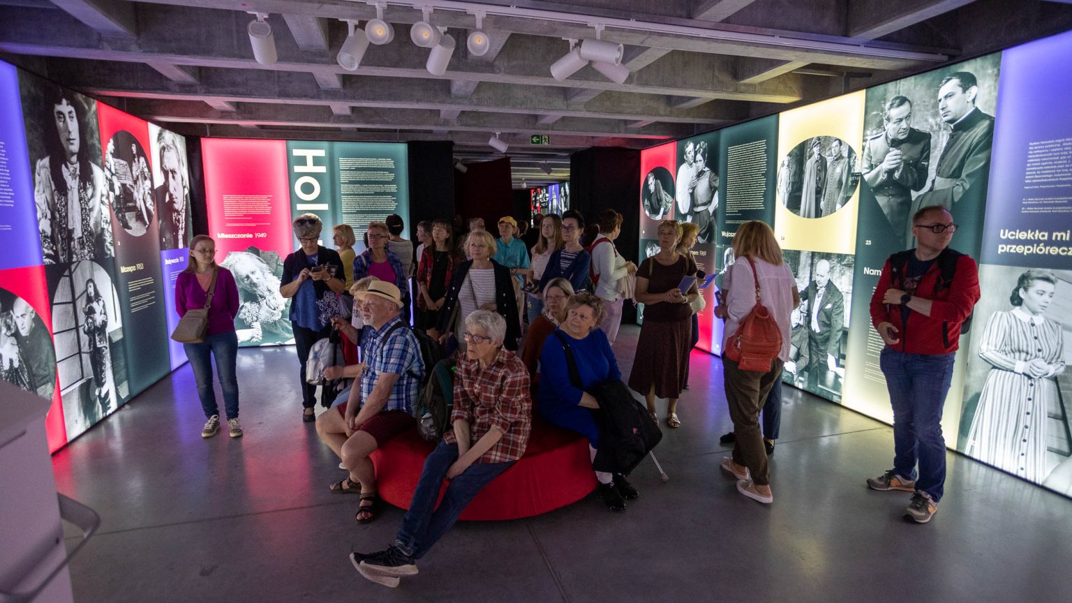 Zwiedzanie wystawy poświęconej artyście w Pałacu Czapskich. Część osób siedzi na pufie po środku sali, na ścianach wokół eksponowane zdjęcia z różnych ról scenicznych Holoubka.