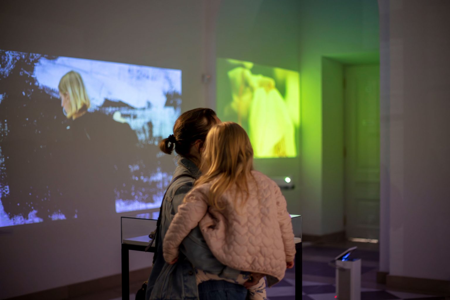 Kobieta z dziewczynką na ręku w sali wystawowej z wyświetlanymi na ścianach obrazami z projektora.