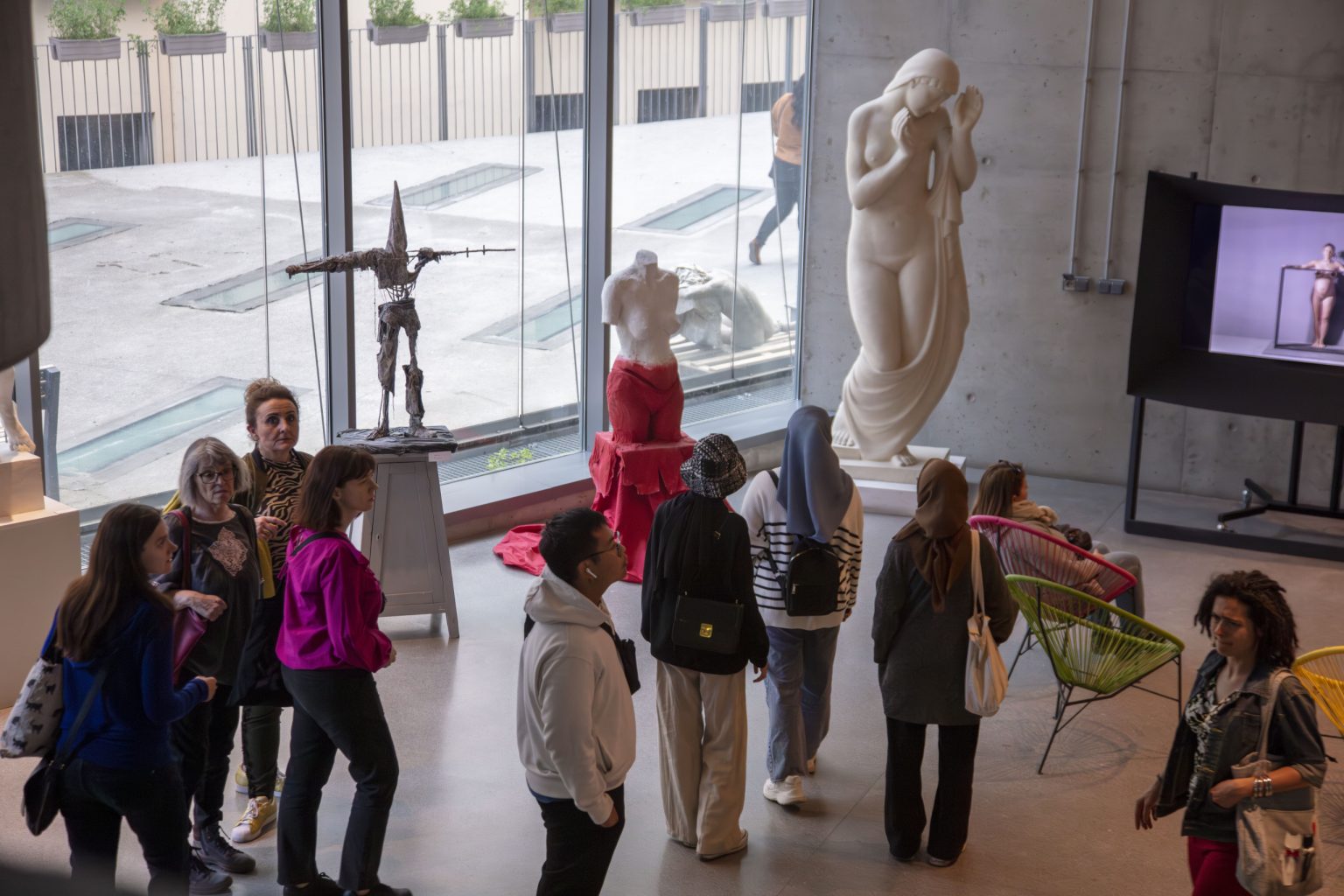Grupa ludzi w sali wystawowej obserwujących rzeźby. Jedna z kobiet siedzi na krześle przed telewizorem