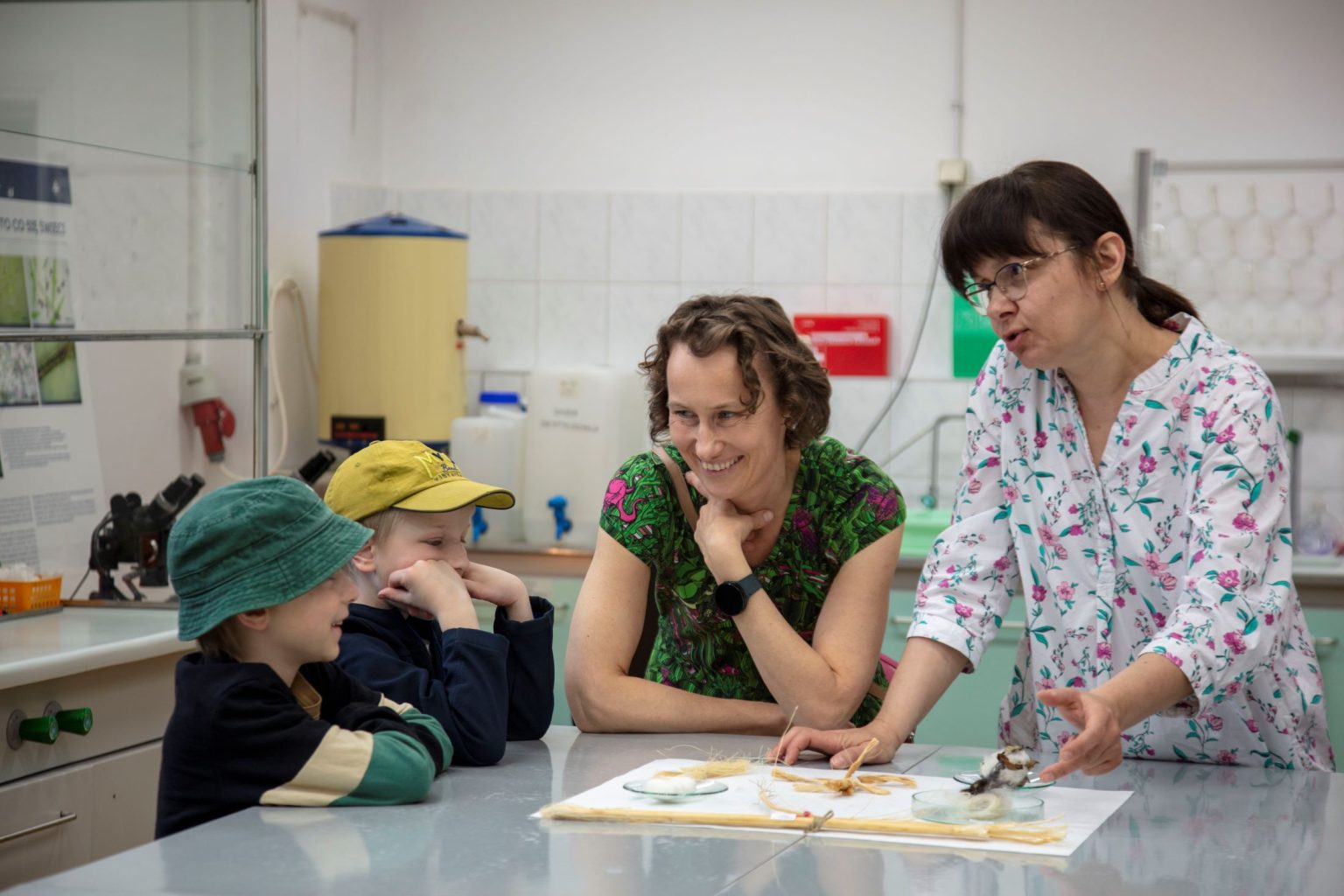 Dwie kobiety przy stole rozmawiają z dziećmi, na blacie na rozłożonej kartce są obiekty przypominające słomę,