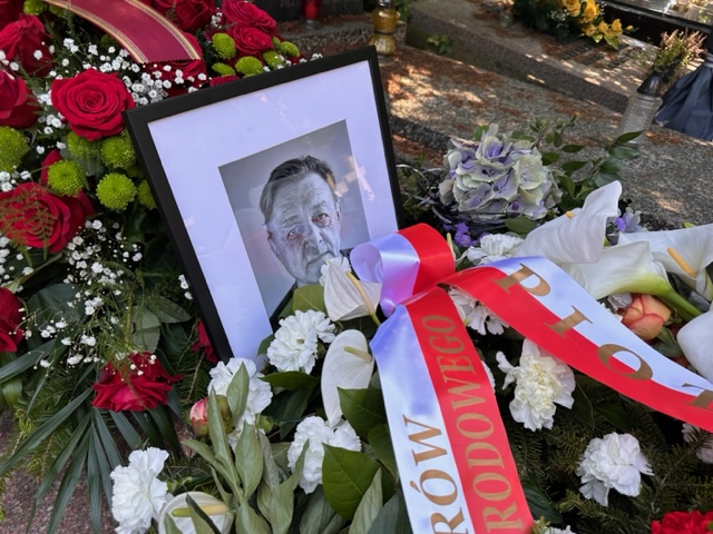 Zdjęcie nagrobka. Wokół czarno-białej fotografii, ułożone są białe kwiaty, przepasane biało-czerwoną szarfą