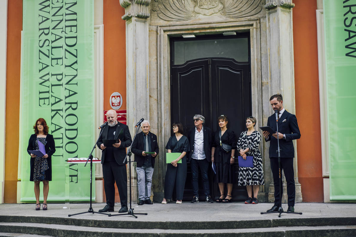 Przemawia Błażej Ostoja - Lniski, w tle jurorzy 28. Międzynarodowego Biennale Plakatu